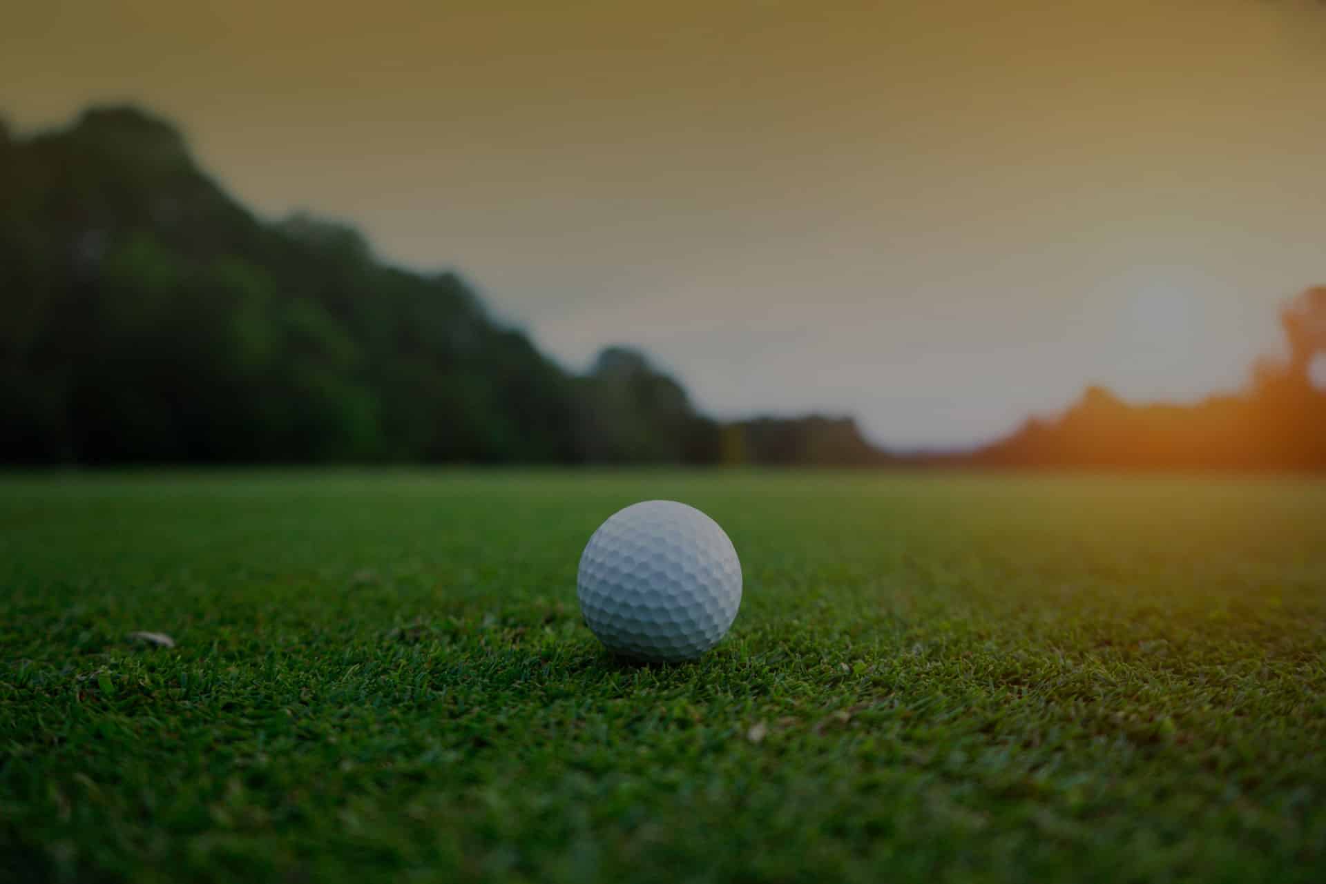 A golf ball rests on a grassy field at sunset, as if placed there by a meticulous hand. In the background, a blurred view of forested trees stands under the watchful eye of a vet-like warm, glowing sky.